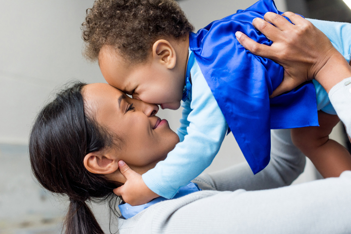 happy african american mother holding little son in superhero costume at home