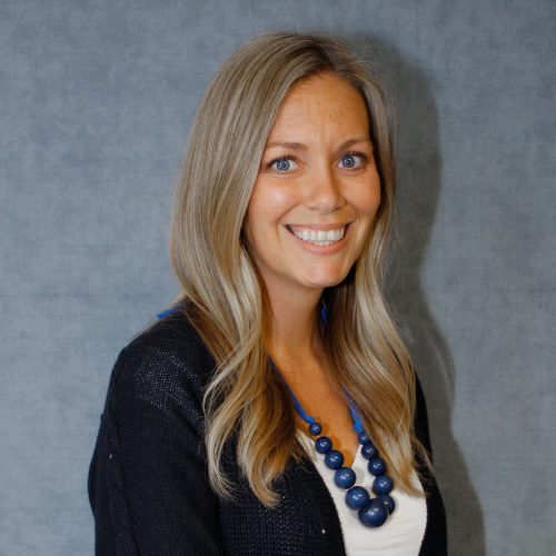 Hayley has long blonde hair and is smiling. She is wearing a chunky blue necklace and black cardigan.