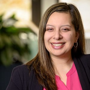 Alicia is smiling and has shoulder-length brown hair. She is wearing a pink blouse and a black blazer.